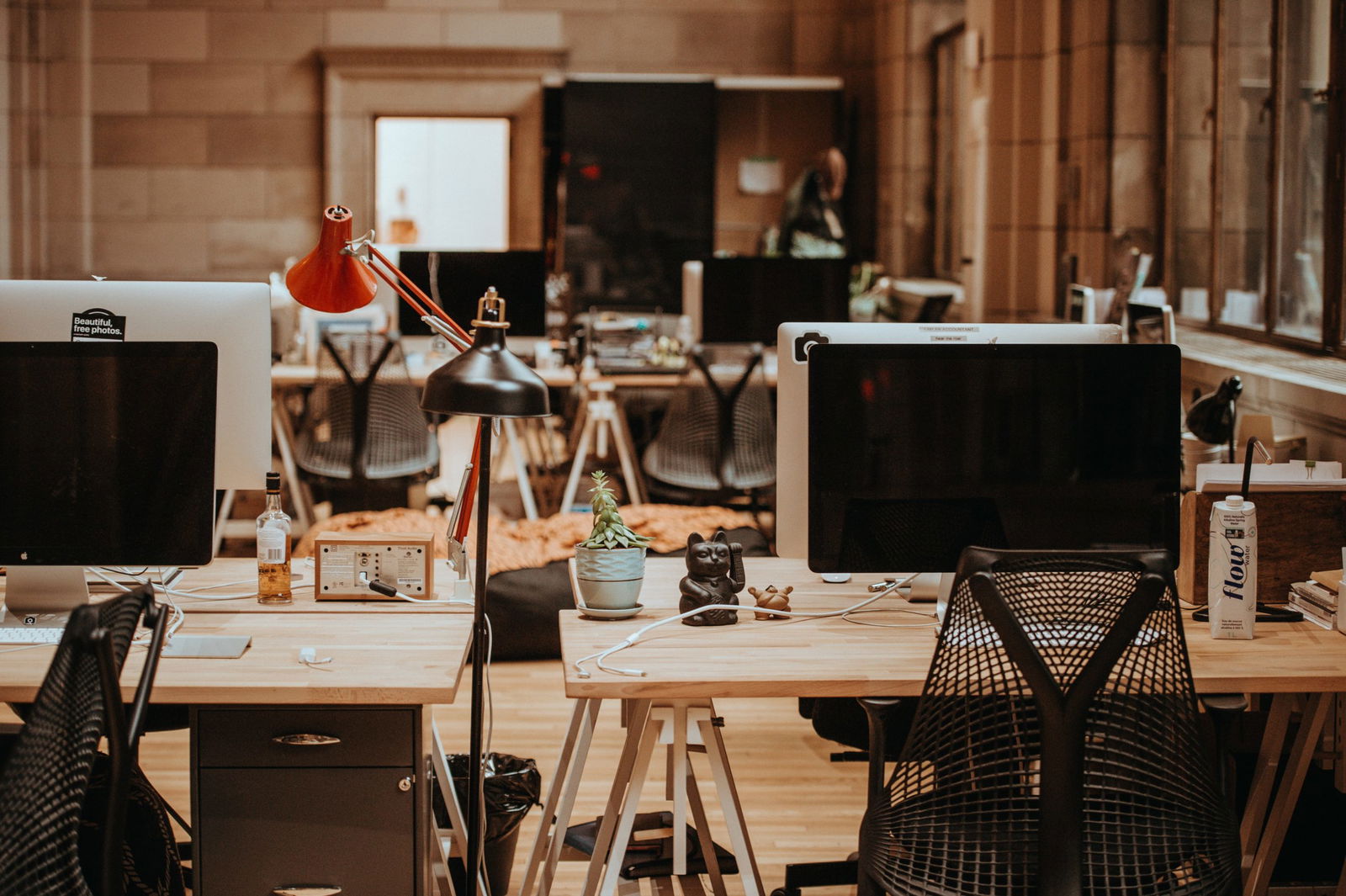 A modern office space with several workstations, computers, and office chairs. A red desk lamp and small plant are visible on the nearest desk. The area appears well-lit and organized.