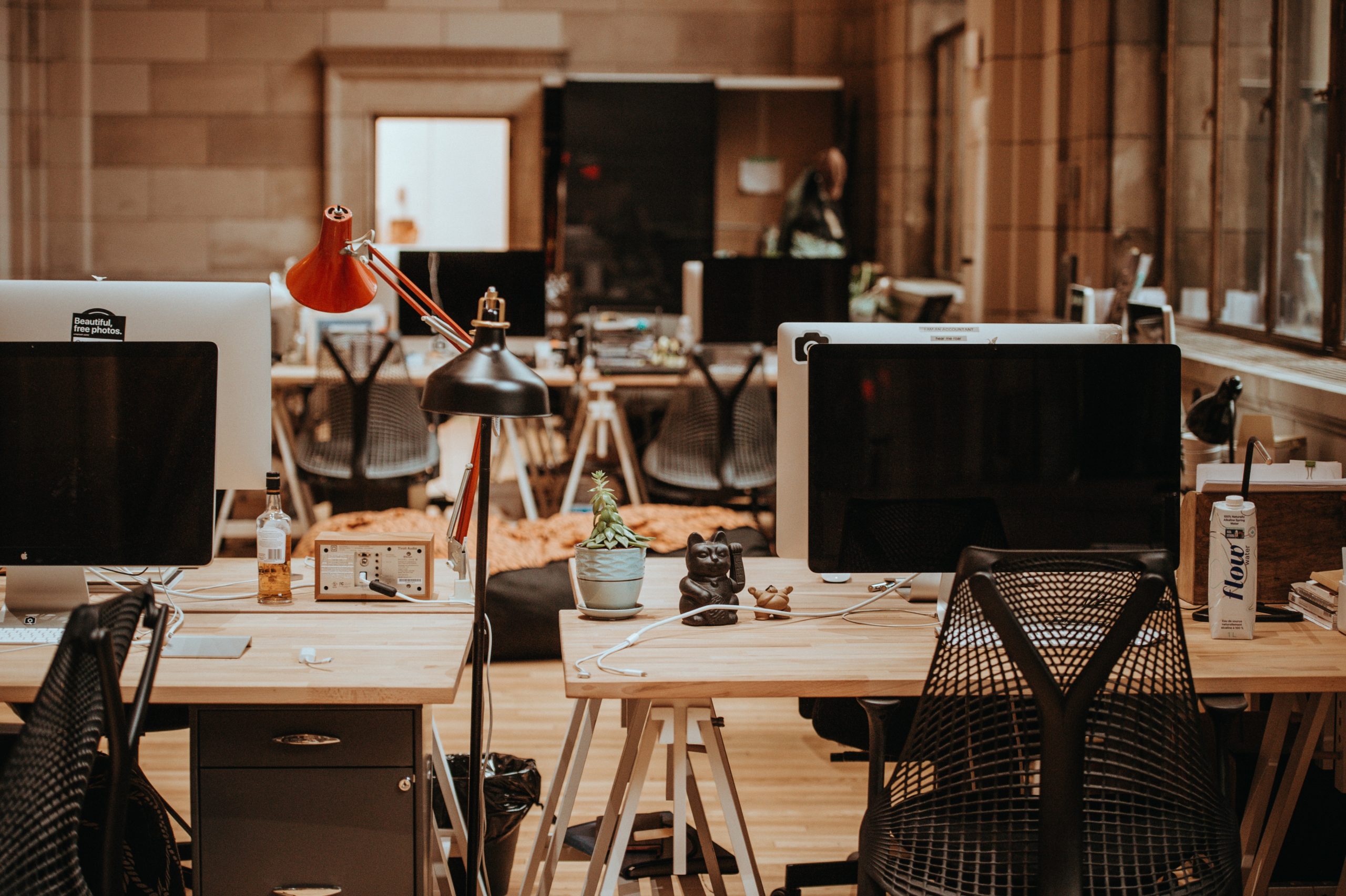 A modern office space with several workstations, computers, and office chairs. A red desk lamp and small plant are visible on the nearest desk. The area appears well-lit and organized.