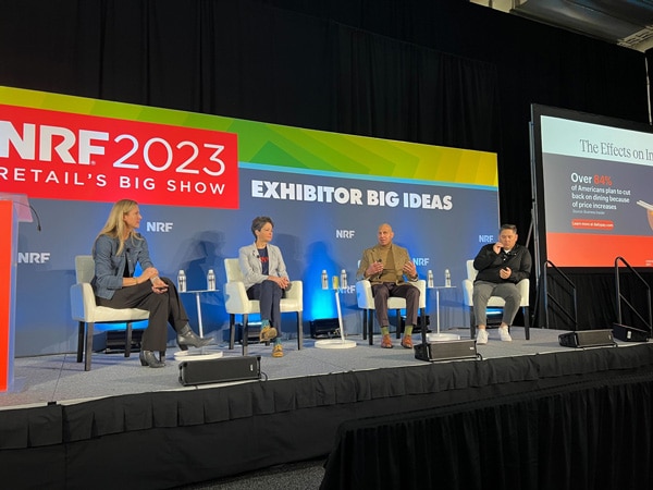 Four speakers sit on a stage during the NRF 2023 event, "Retail's Big Show," with a screen displaying a presentation slide titled, "The Effects on Retail.