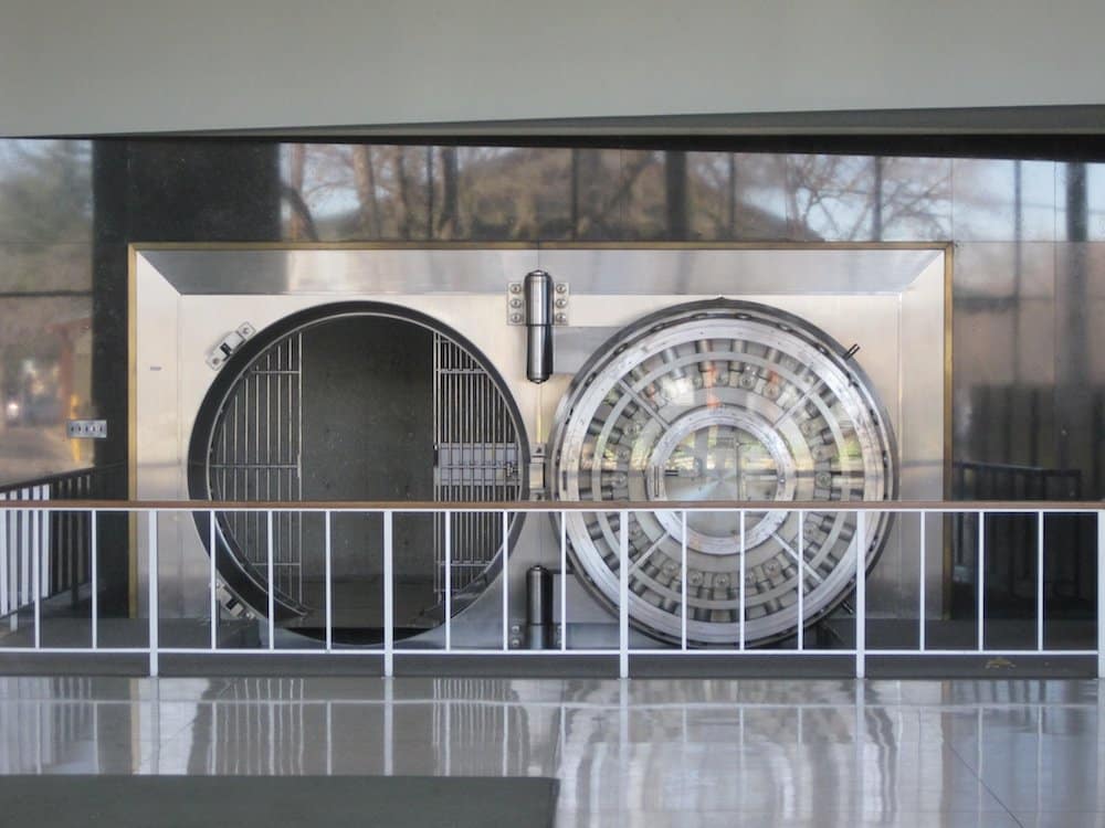A large, circular, metal vault door is open, revealing the entrance to a secure room with a steel gate. The area is enclosed by a white railing—a perfect place to save more money securely.