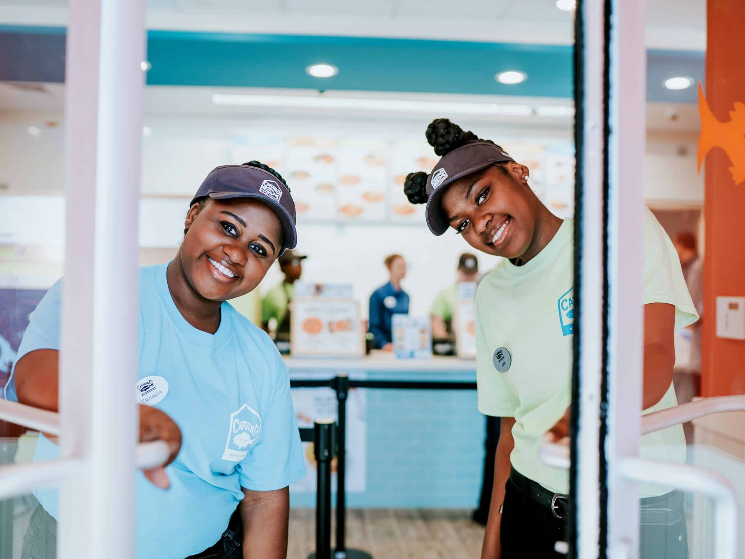 Two women wearing uniforms and caps smile while standing at the entrance of a store. The interior of the store and other staff members are visible in the background.