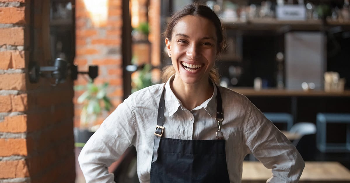 A smiling woman wearing a black apron stands inside a brick-walled establishment, possibly a cafe or restaurant.