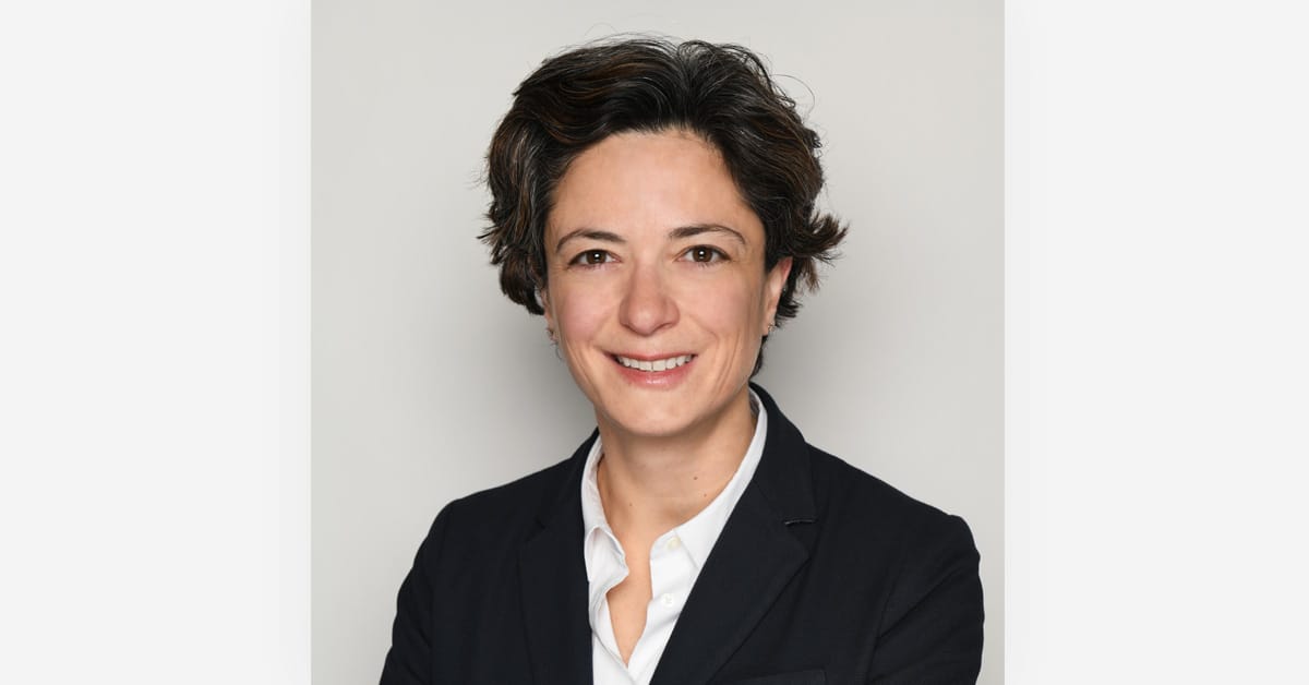 A person with short dark hair wearing a black blazer and white shirt smiles at the camera against a plain background.