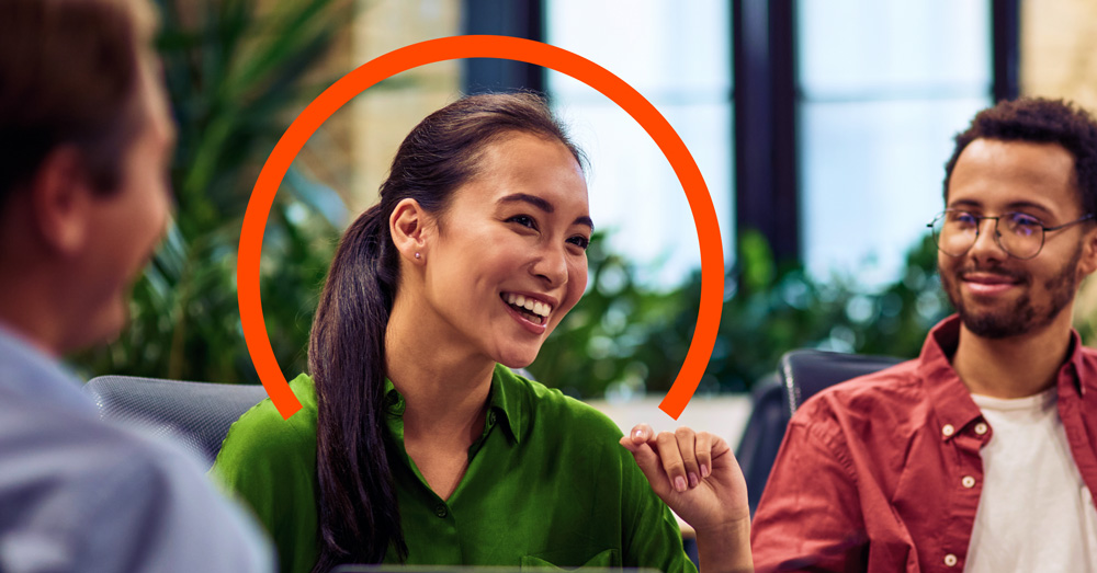 A woman in a green shirt smiles during a group meeting, with an orange semicircle graphic behind her head. Two other people are seated nearby.