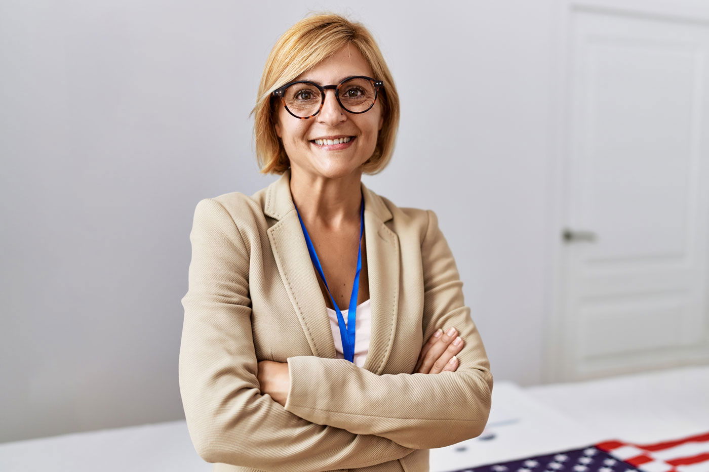 A woman with glasses and a beige blazer stands confidently with her arms crossed in front of a table with a partial view of an American flag.