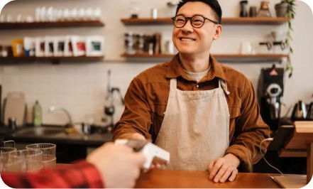 A barista wearing glasses and a brown shirt with an apron smiles while handing a coffee cup to a customer at a café counter.