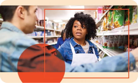 A woman wearing a white apron stands in a grocery store aisle, looking at a person who is talking to her. She has one hand on a shelf with various items.