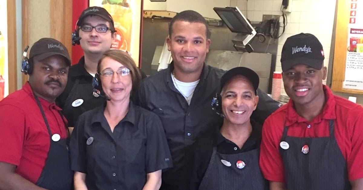 Six employees wearing Wendy's uniforms stand together and smile in a Wendy's restaurant kitchen.