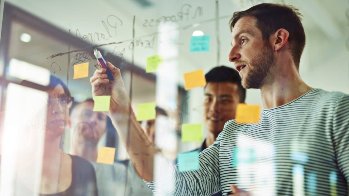 A man writes on a glass board with sticky notes as three others look on, appearing to be in a collaborative meeting or brainstorming session.