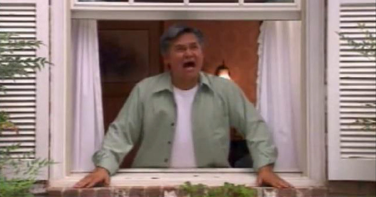 A man with gray hair and wearing a light green shirt is standing at a window, looking outside and shouting.
