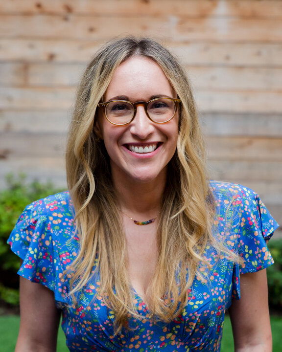A woman with long blonde hair and glasses smiles at the camera, wearing a blue floral dress, standing outdoors in front of a wooden wall.