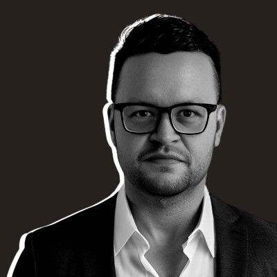 A man with short, dark hair, beard, and glasses wears a white shirt and dark blazer, standing against a dark background.