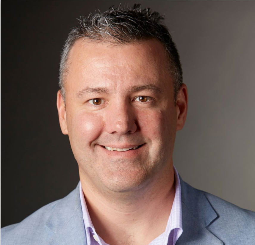 A man with short gray hair and a light beard, wearing a light blue blazer and light purple shirt, smiling at the camera against a plain background.