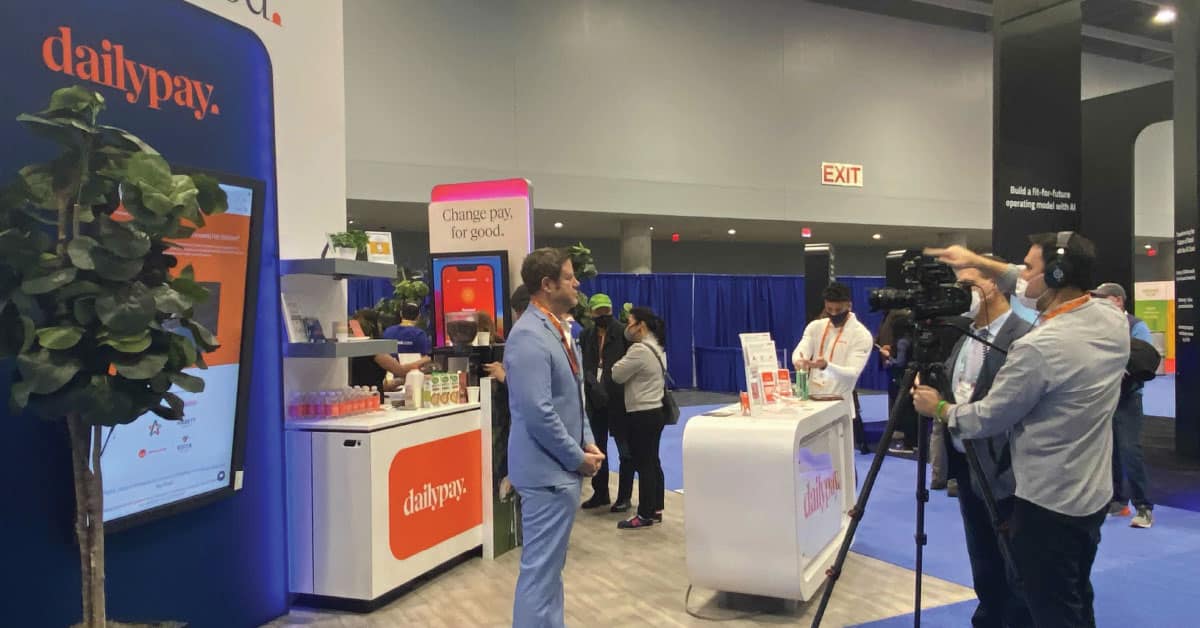 A man in a suit is being filmed by a camera crew at a booth labeled “dailypay” at an indoor exhibition. The booth has a large screen and promotional materials displayed. Other people are in the vicinity.