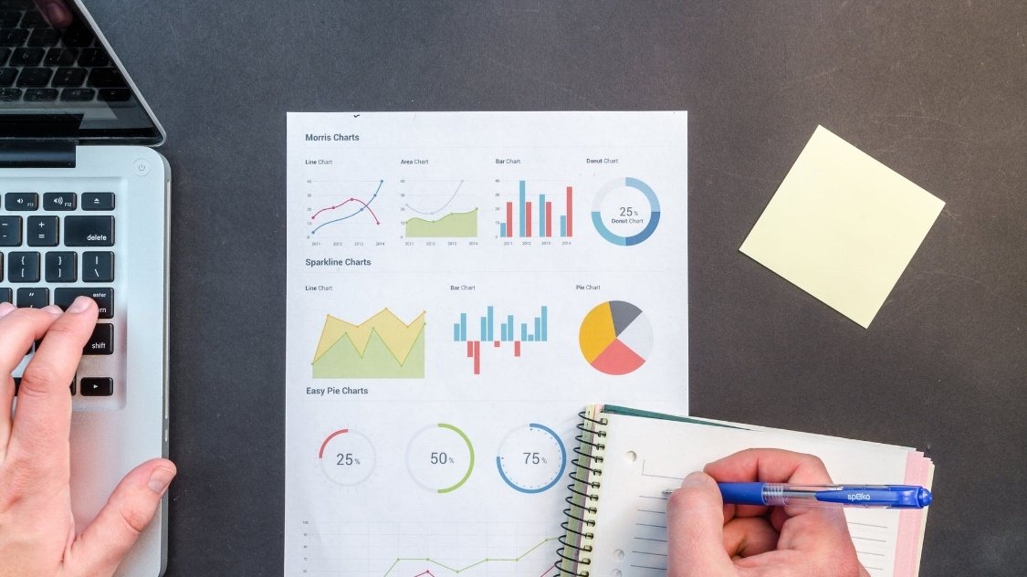 A person types on a laptop with their left hand and writes in a notebook with their right. A sheet with various colorful charts and a yellow sticky note are on the table.