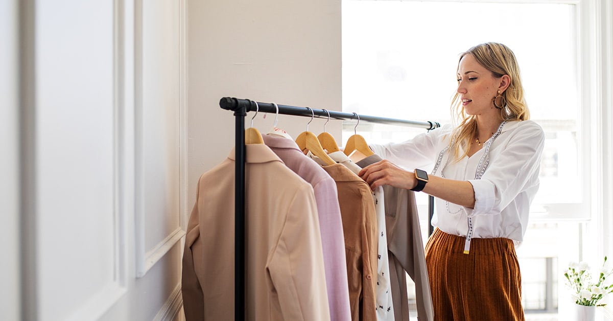 A woman in a white blouse sorts through clothing on a rack with several items hanging on wooden hangers in a well-lit room.