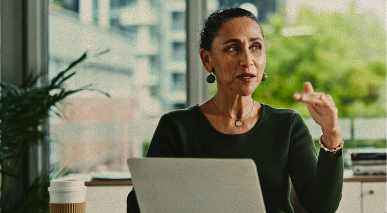 A woman sits at a desk with a laptop, gesturing as she speaks. There is a coffee cup nearby and large windows with a view of trees and buildings in the background.
