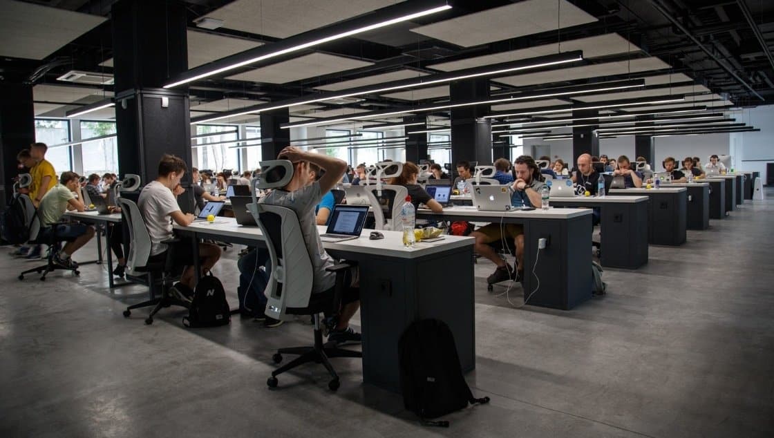A large, modern office with rows of people working on laptops at long desks, with overhead lighting and a minimalist design.