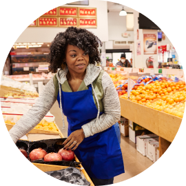 A person wearing a blue apron and a sweater is arranging produce items in a grocery store.