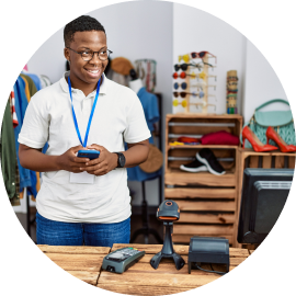 A person in a retail store stands behind a checkout counter with a barcode scanner, card reader, and monitor. They are holding a phone, smiling, and wearing a blue lanyard.