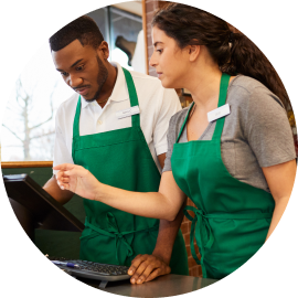 Two employees wearing green aprons work at a cash register in a store. One points at the screen while the other looks on attentively.