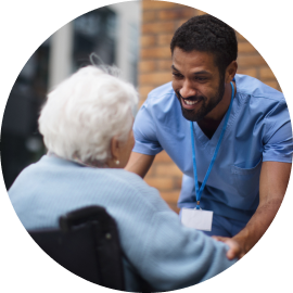 A healthcare worker in blue scrubs smiles and leans towards an elderly person in a wheelchair during a conversation outdoors.