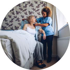 A caregiver assists an elderly woman in putting on a shirt as she sits on the edge of a bed in a bedroom.