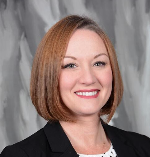 A woman with straight, chin-length light brown hair wearing a black blazer and white patterned top, smiling in front of a gray and white background.