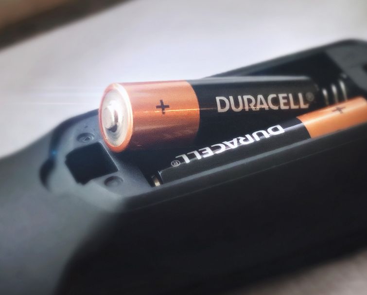 Close-up of two Duracell AA batteries inside a black electronic device, with one battery partially inserted and the other fully in place.