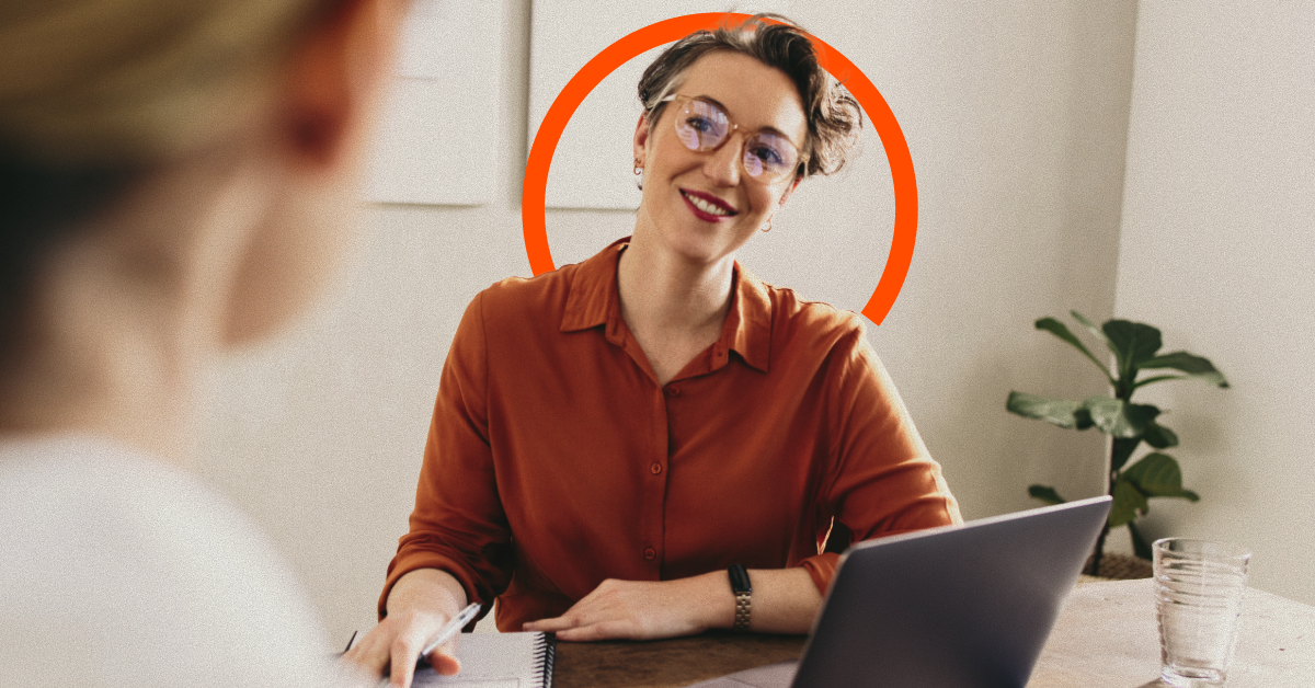 A person in glasses and an orange shirt smiles while sitting at a desk with a laptop, speaking to another person. A circular orange design is behind their head.
