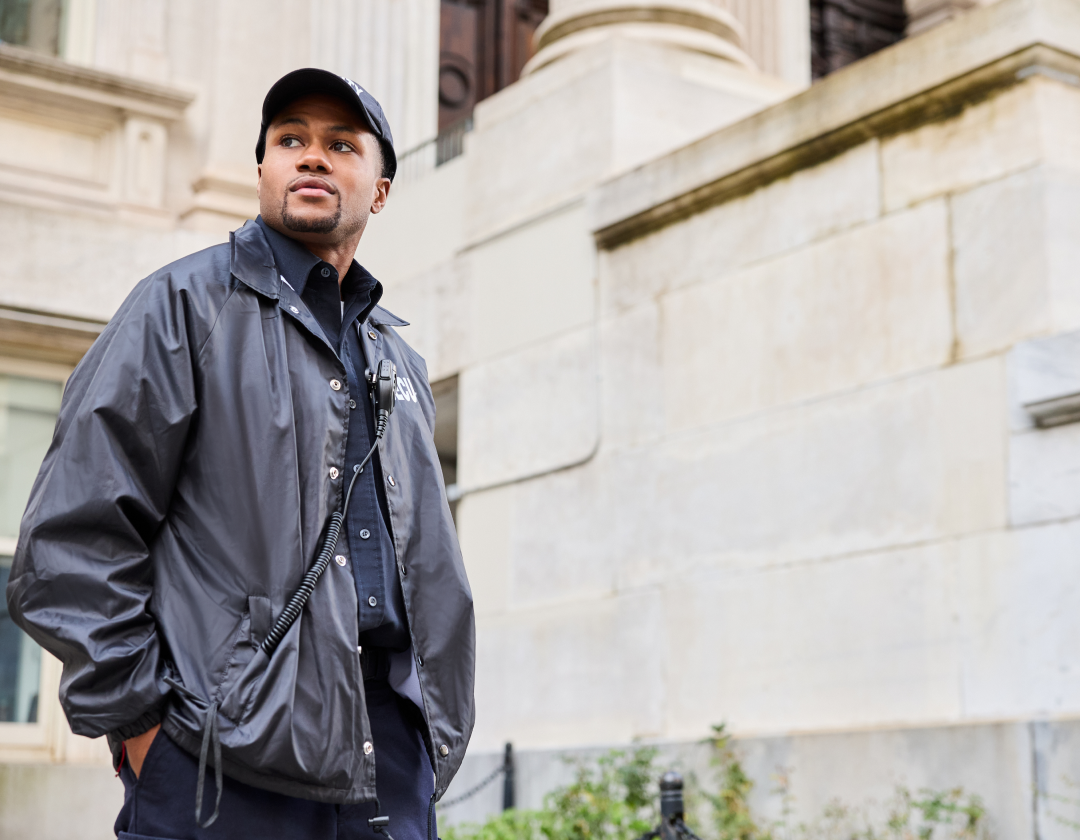 A uniformed security guard stands outside a stone building, looking to the side with one hand in his pocket.