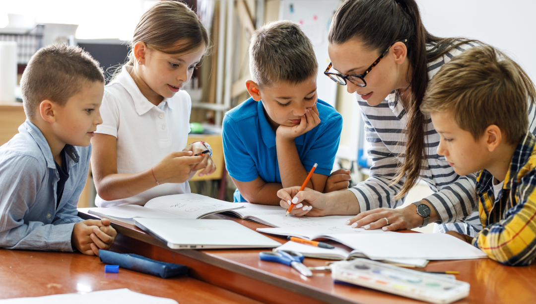 A woman helps four children with their schoolwork at a table, surrounded by open books and stationery.