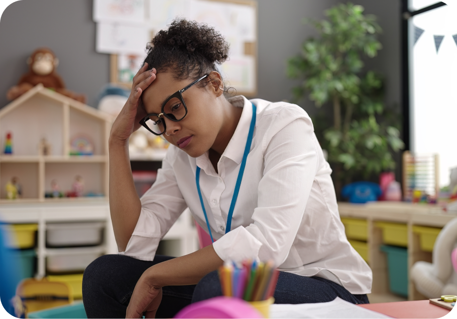 A woman in casual office attire and glasses sits in a room with children's toys, resting her forehead on her hand, appearing stressed or deep in thought.
