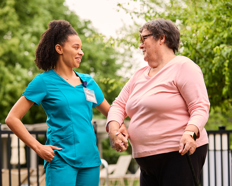 A healthcare worker in blue scrubs smiles and holds hands with an older woman in a pink top outdoors, suggesting support or assistance.
