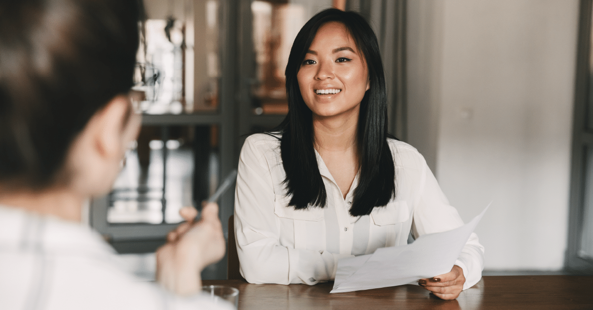 A woman with long dark hair and a white blouse sits at a wooden table, smiling and holding a sheet of paper. She is engaged in a conversation with another person, who is slightly out of focus in the foreground and appears to be holding a pen. The background features glass doors and a modern interior.