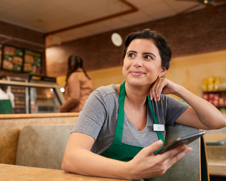 A woman wearing a green apron sits at a restaurant table, holding a phone and smiling, with another person in the background near the counter.