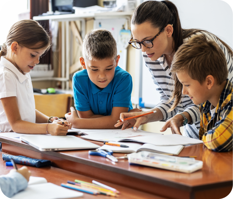 A woman guides three children working on assignments at a classroom table, with notebooks and stationery spread out in front of them.