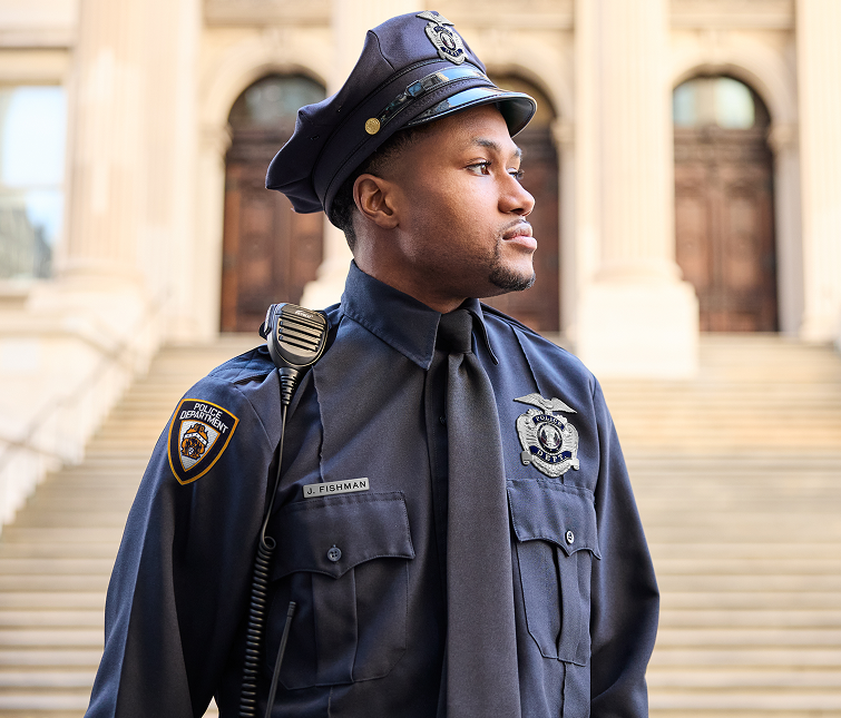 A police officer in uniform stands outside a building with stone steps and columns, looking to the side.