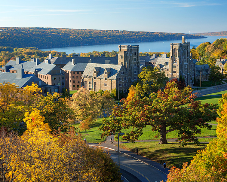 A university campus with stone buildings, surrounded by autumn trees, overlooking a large lake under a clear blue sky.