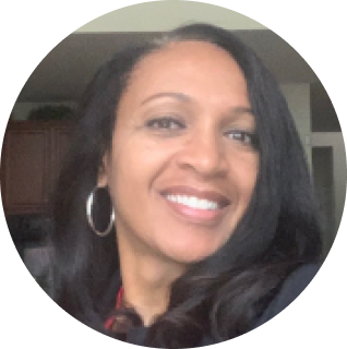 A woman with long black hair and hoop earrings smiles at the camera in an indoor setting.