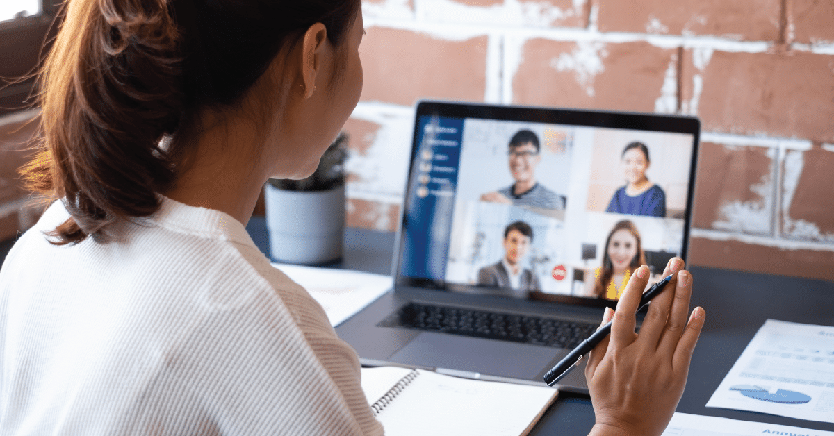 A person sits at a desk participating in a video conference on a laptop. They hold a pen and a notebook is open in front of them. Four people are visible on the laptop screen.