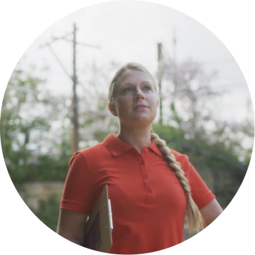 A person in a red polo shirt stands outdoors, holding a tablet, with trees and power lines in the background.